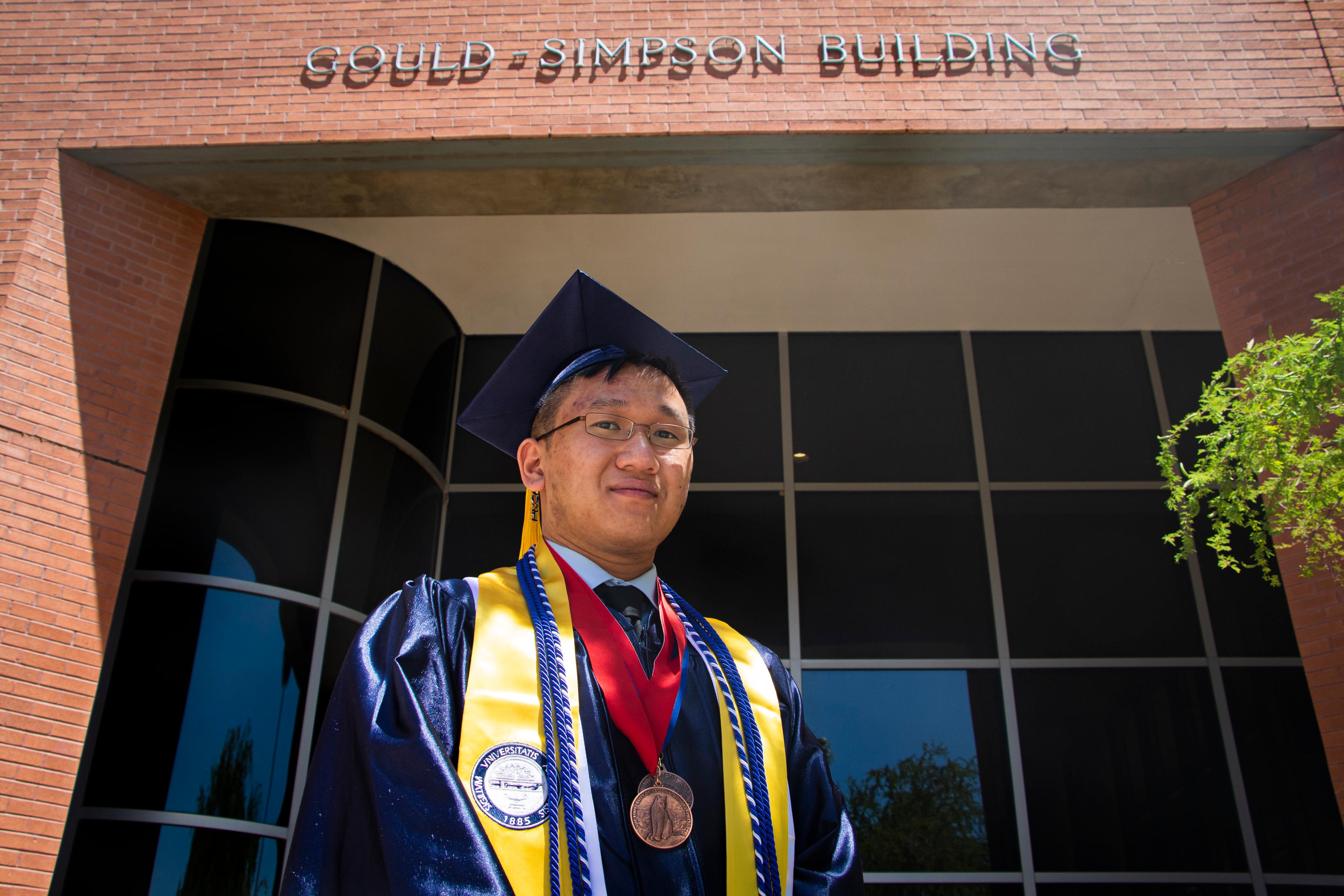Ryan in cap and gown with honors cords and medals in front of the University of Arizona Gould-Simpson Building