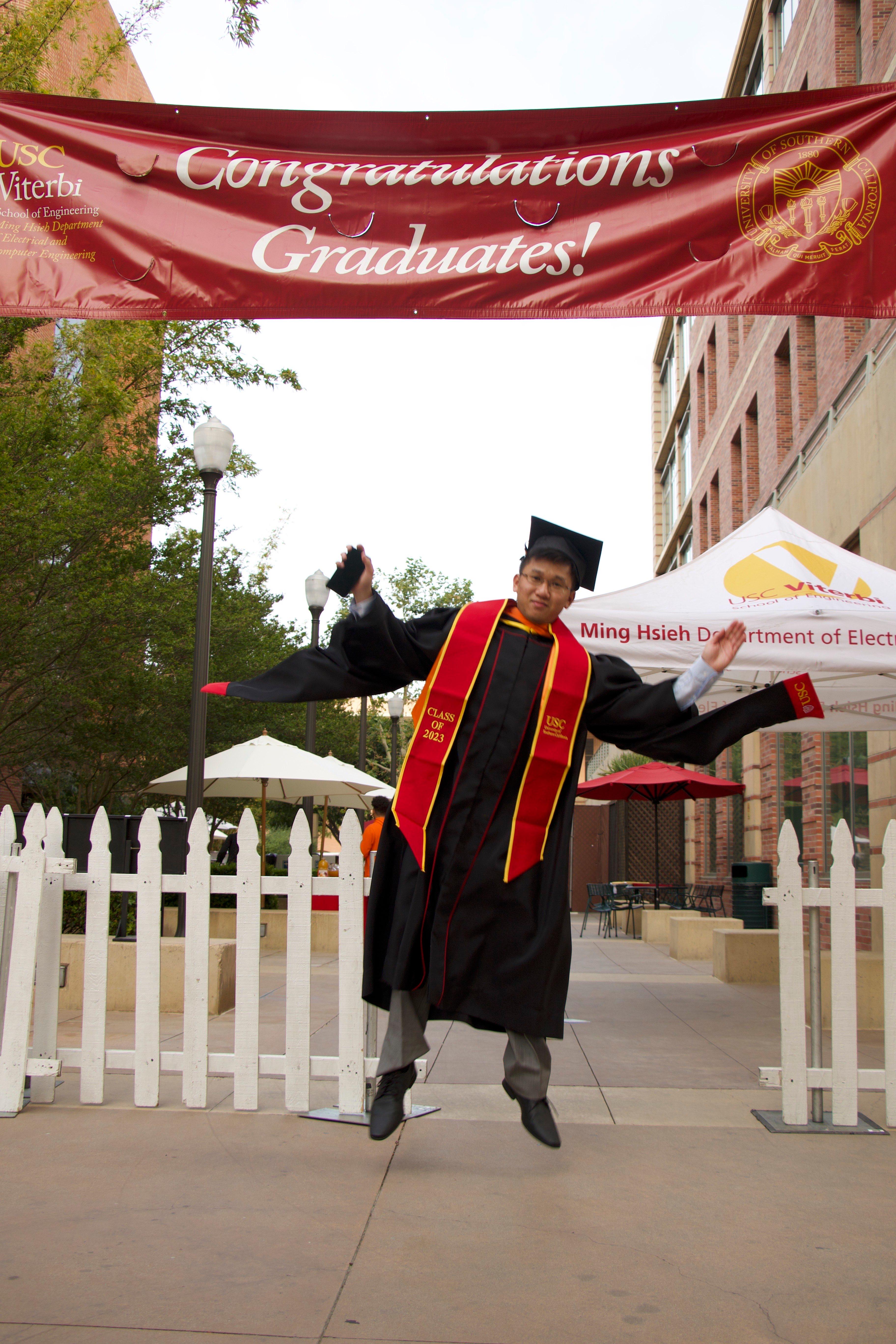 Ryan jumping in his USC cap and gown with a Class of 2023 stole beneath a USC Viterbi Congratulations Graduates banner