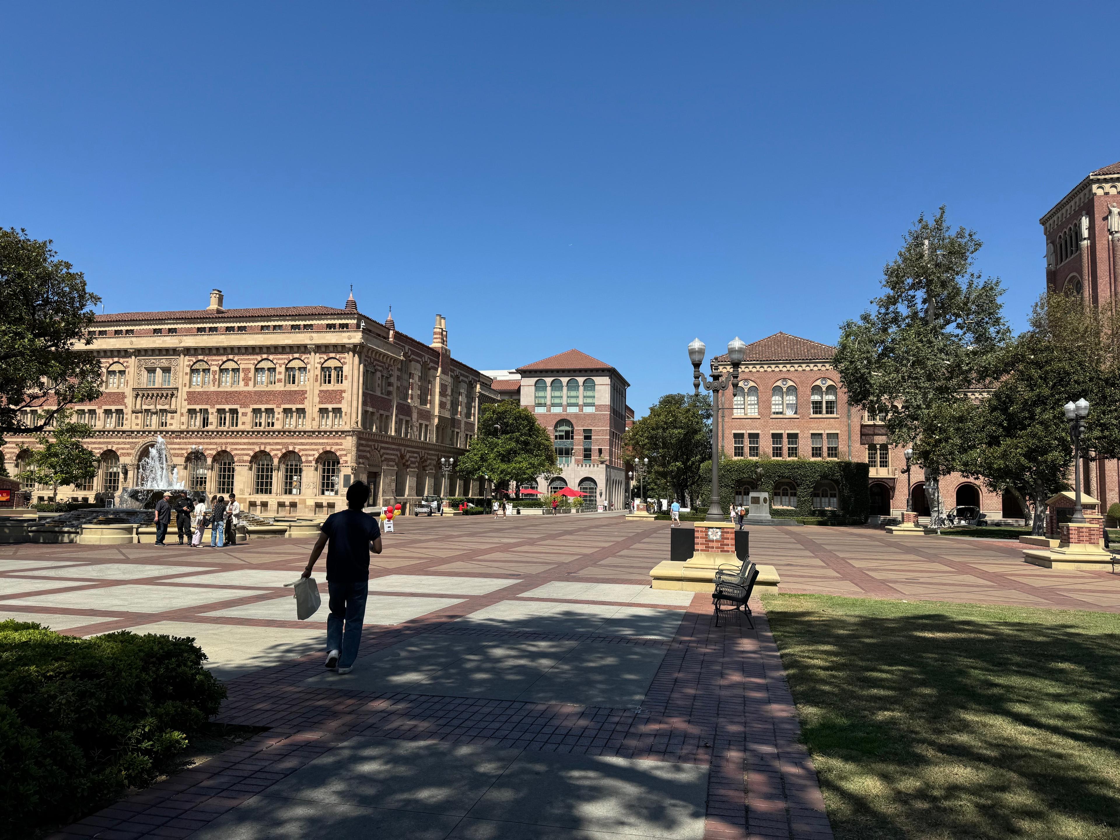 A person walking across USC's Hahn Plaza past the fountain and brick buildings under a clear blue sky